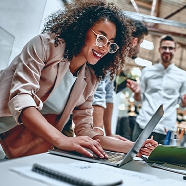 Smiling businesswoman typing on a laptop at a collaborative workspace, with colleagues conversing in the background.