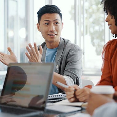 Professional man speaking and gesturing during a team meeting in a modern office, with laptops and coffee cups on the table.