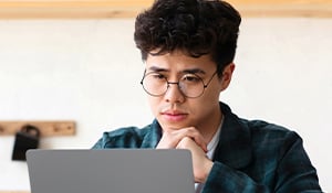 Young person wearing glasses sitting in a kitchen, looking perplexed at a laptop screen while resting their chin on their hands.