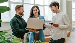 Three coworkers standing together in a bright, plant-filled office, reviewing content on a laptop while discussing ideas.