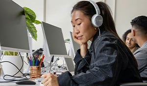 A woman wearing headphones works at a desk in an open office, writing in a notebook beside a computer, with coworkers and monitors in the background.