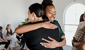Two women warmly hugging in the foreground, while a small group of people sit and talk together in the background against a light-colored wall.