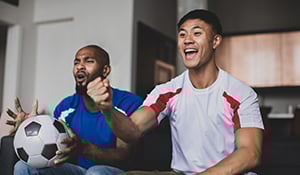 Two men sitting on a couch excitedly watching a soccer game, one holding a soccer ball while both cheer and smile.
