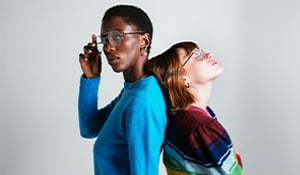 Two people standing back-to-back. One person in a bright blue sweater looks directly at the camera while adjusting their glasses. The other, in a colorful striped sweater, leans back with their head tilted upward and eyes closed, creating a contrast in posture and mood.