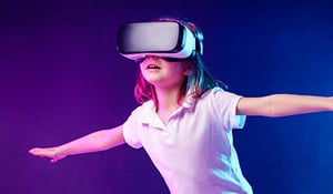 Young girl wearing a virtual reality (VR) headset, arms outstretched as if balancing or flying, immersed in a digital experience against a gradient purple and blue background.