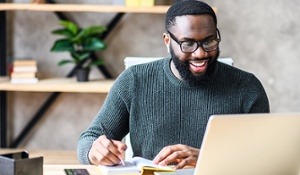 African American man looks at his laptop while actively writing in a notebook