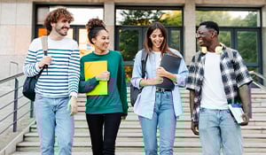 Four smiling students walk down campus steps, chatting and carrying books and backpacks.
