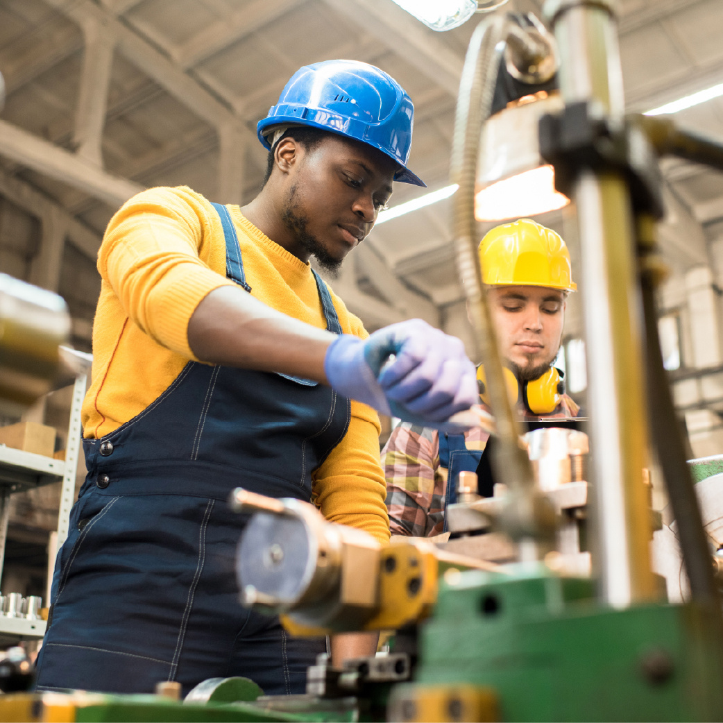 Two industrial workers wearing protective helmets and gloves operate machinery in a manufacturing facility.