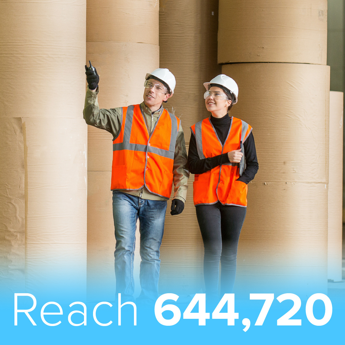 Two workers wearing white hard hats and orange safety vests walk through a paper manufacturing facility, surrounded by large rolls of paper. One worker gestures while speaking, and the other listens attentively. A blue banner at the bottom displays the text 