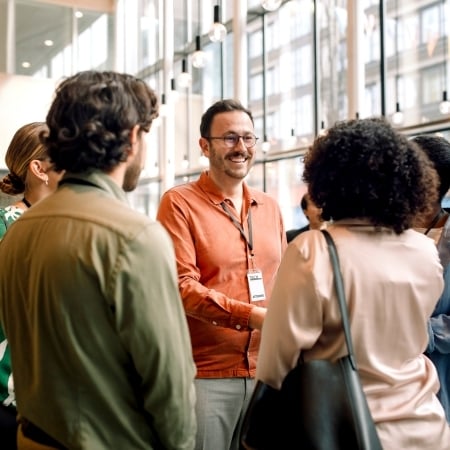 A group of professionally dressed people engaged in conversation at a networking event or conference. One man in an orange shirt and lanyard is smiling and shaking hands with a woman, with large windows in the background letting in natural light.