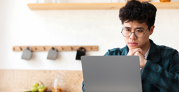 Young person wearing glasses sitting in a kitchen, looking perplexed at a laptop screen while resting their chin on their hands.