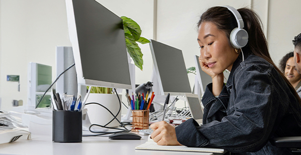 A woman wearing headphones works at a desk in an open office, writing in a notebook beside a computer, with coworkers and monitors in the background.