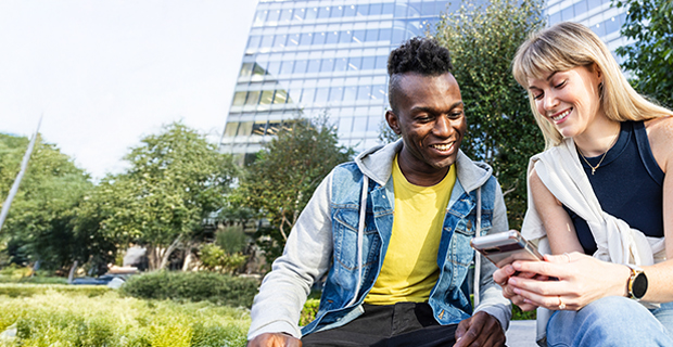 Two young adults sitting outdoors near modern office buildings, smiling and looking at a smartphone together. One wears a yellow shirt and denim jacket, the other a sleeveless top with a white sweater over their shoulders.