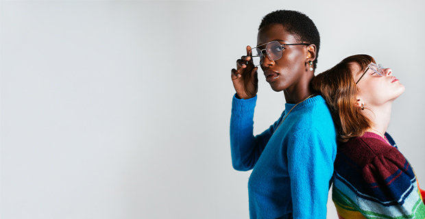 Two people standing back-to-back. One person in a bright blue sweater looks directly at the camera while adjusting their glasses. The other, in a colorful striped sweater, leans back with their head tilted upward and eyes closed, creating a contrast in posture and mood.