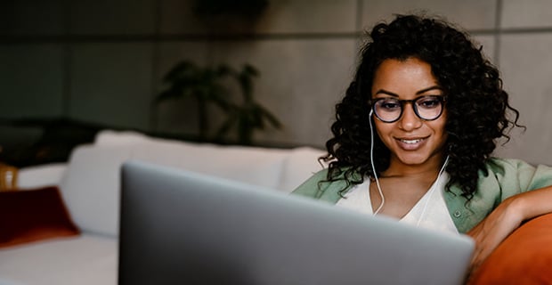 A woman with curly hair and glasses is sitting on a couch, smiling while looking at her laptop. She is wearing earphones and appears to be engaged in a virtual meeting or video call in a modern, relaxed indoor setting.