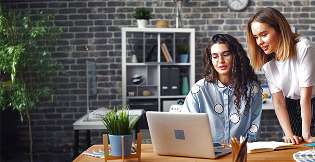 Two women working together at a laptop in a modern office with a brick wall and potted plant.